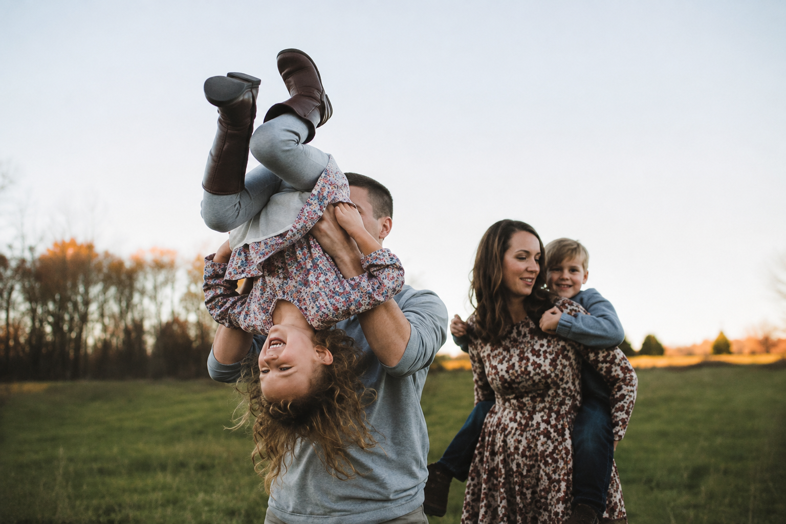 A man holds a laughing girl upside down while an estate planning attorney gives a piggyback ride to a smiling boy in an open grassy field at sunset.