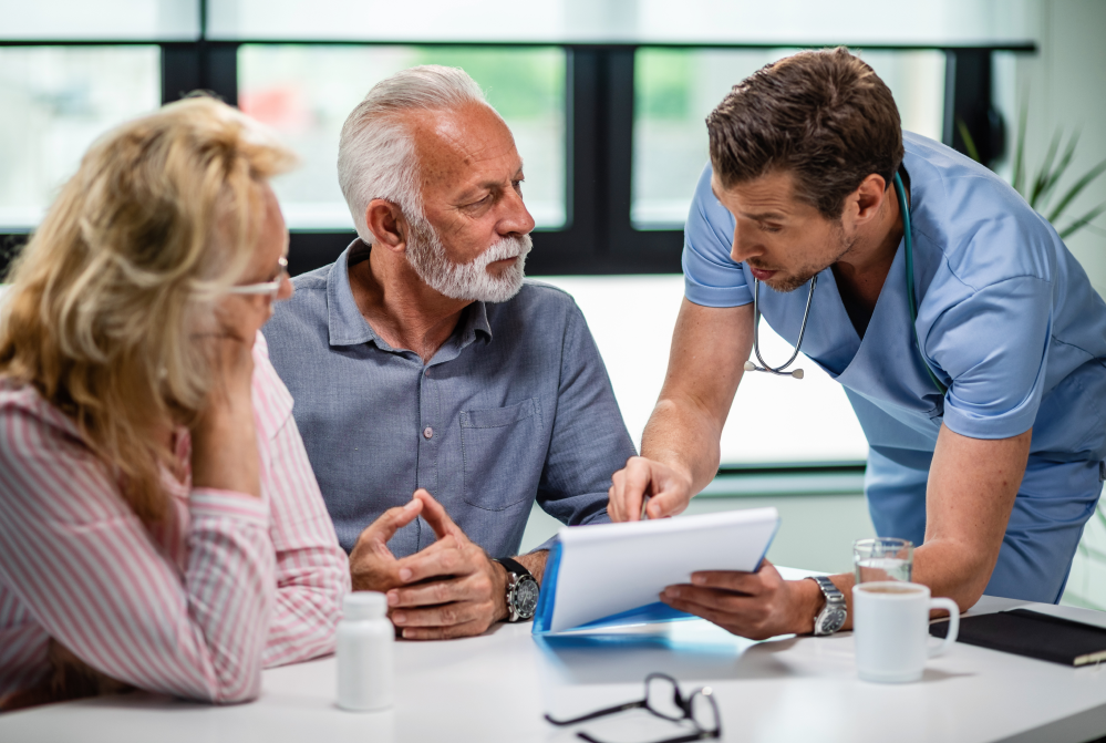 A doctor in scrubs shows documents to an older couple at a table with medication, discussing medical info and Medicaid & Nursing Home Planning Attorney options.