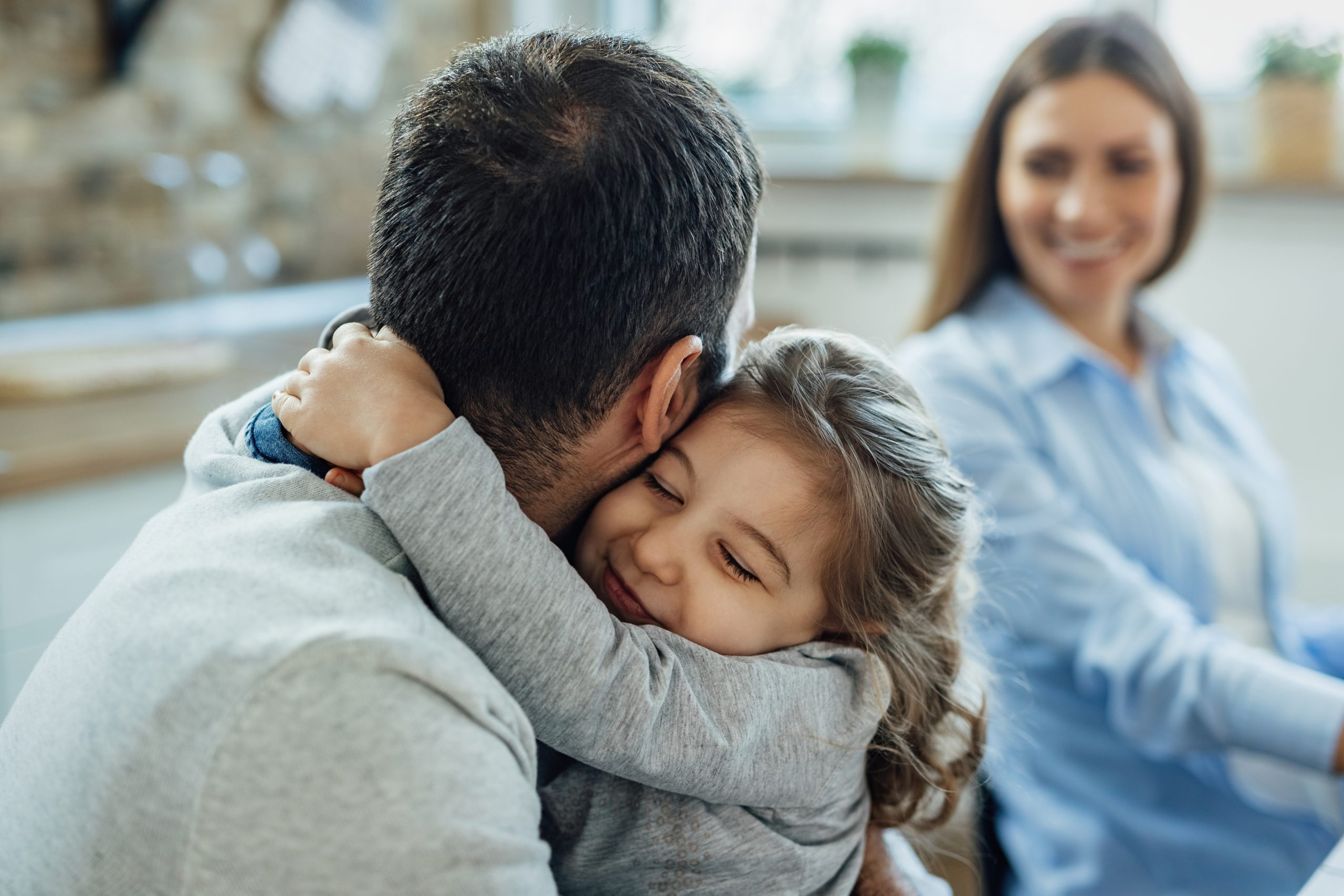 A young girl hugs an adult man while a woman, possibly a guardianship attorney, sits in the background, smiling and watching them.