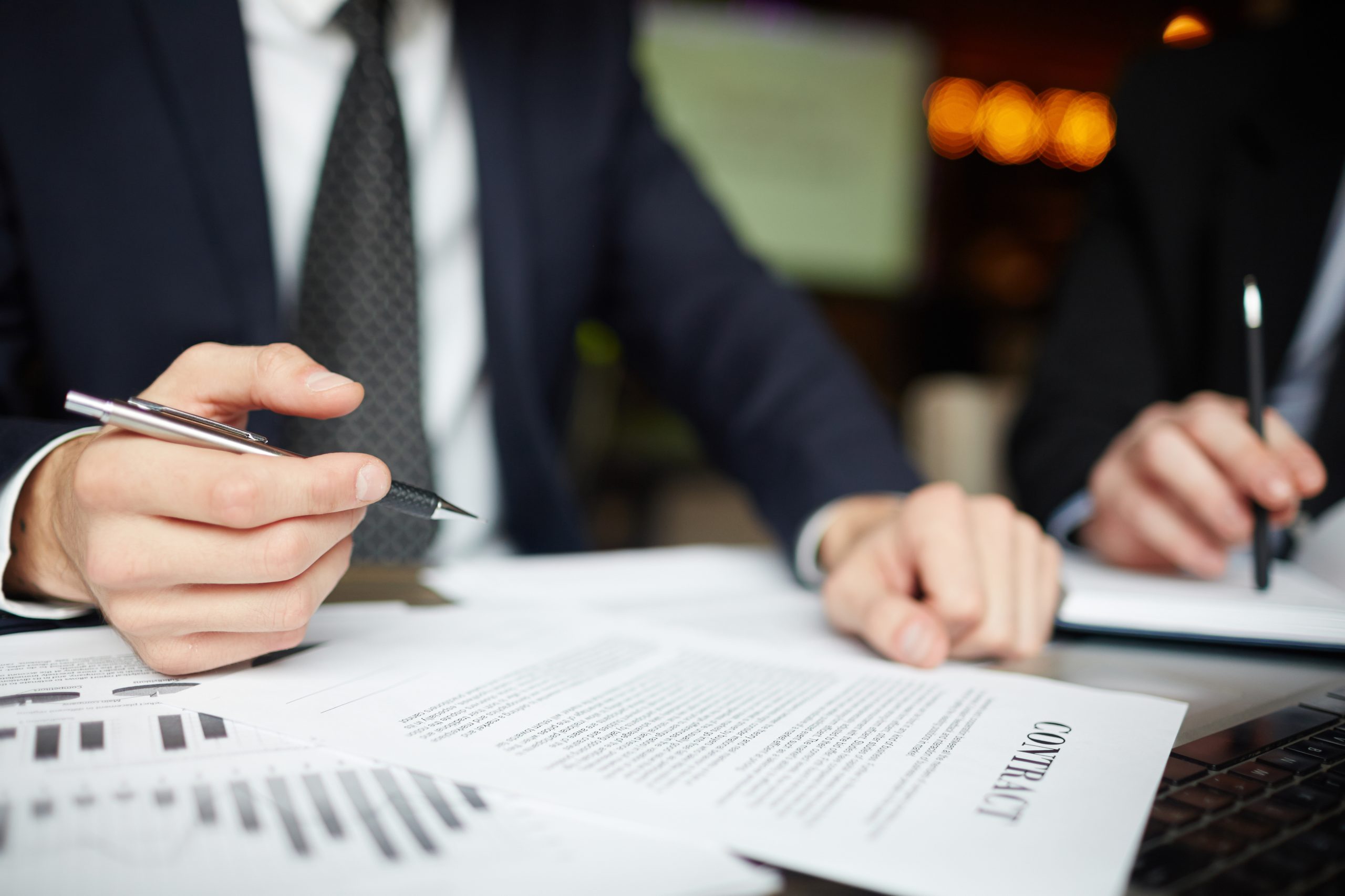 Two people in business suits review documents and charts at a table; one, possibly a Power of Attorney Lawyer, is holding a pen and pointing at a contract.
