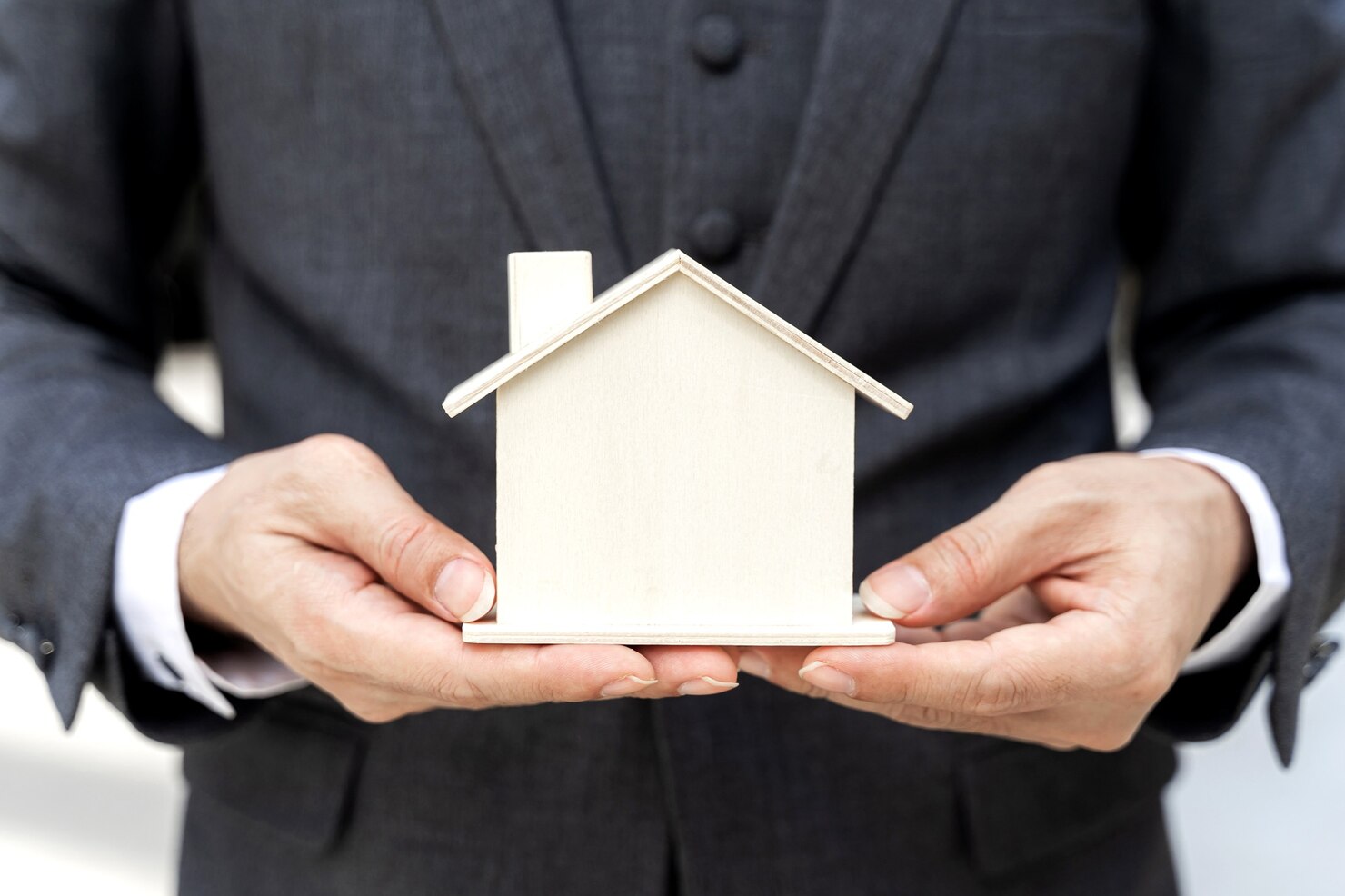 Person in a suit holding a small wooden house model with both hands, symbolizing real estate, home ownership, or guidance from an Asset Protection Attorney.