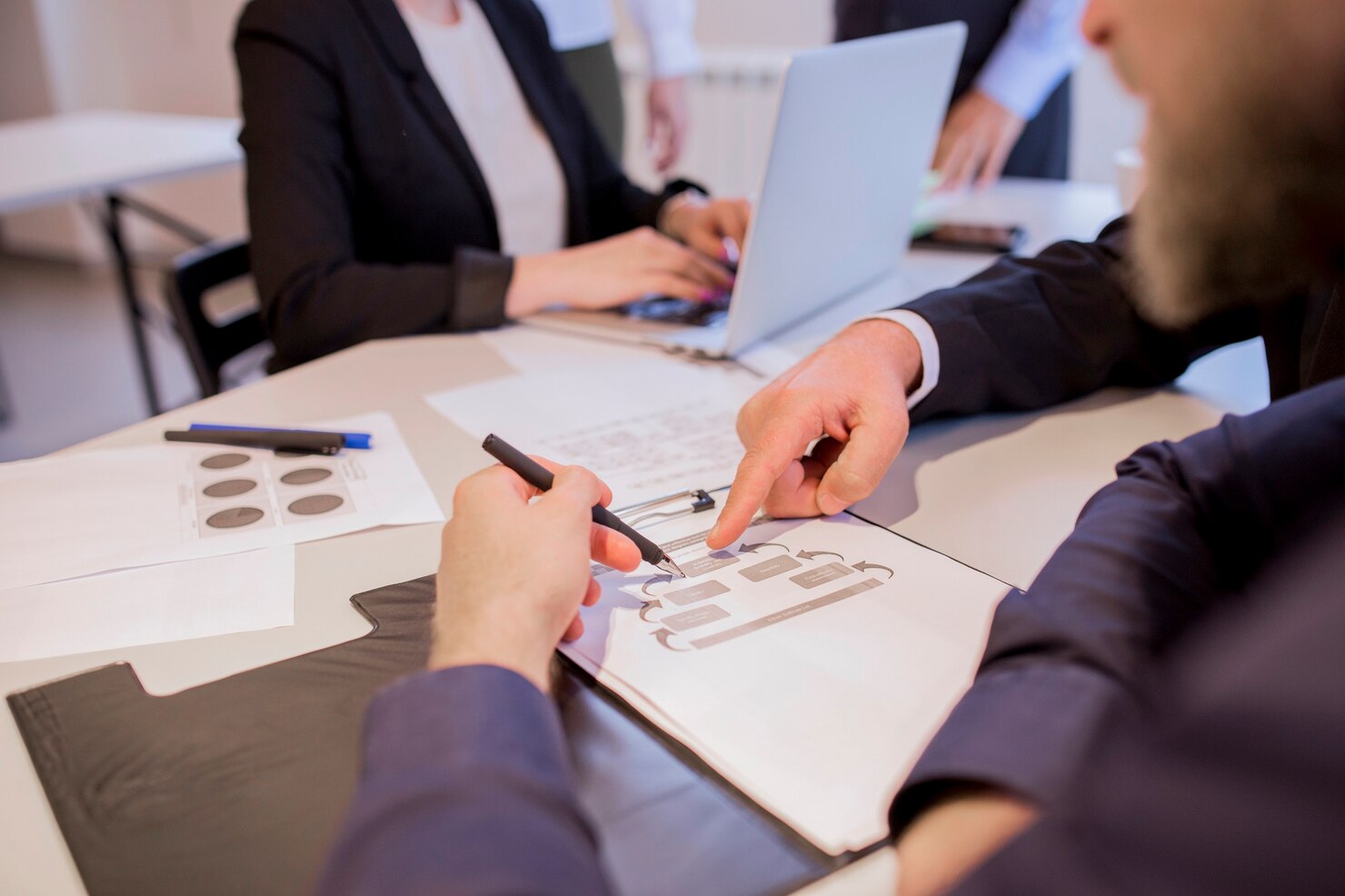 Two people in business attire discuss a flowchart at a table, one pointing at the paper while an Estate Administration Attorney types on a laptop among scattered documents and pens.