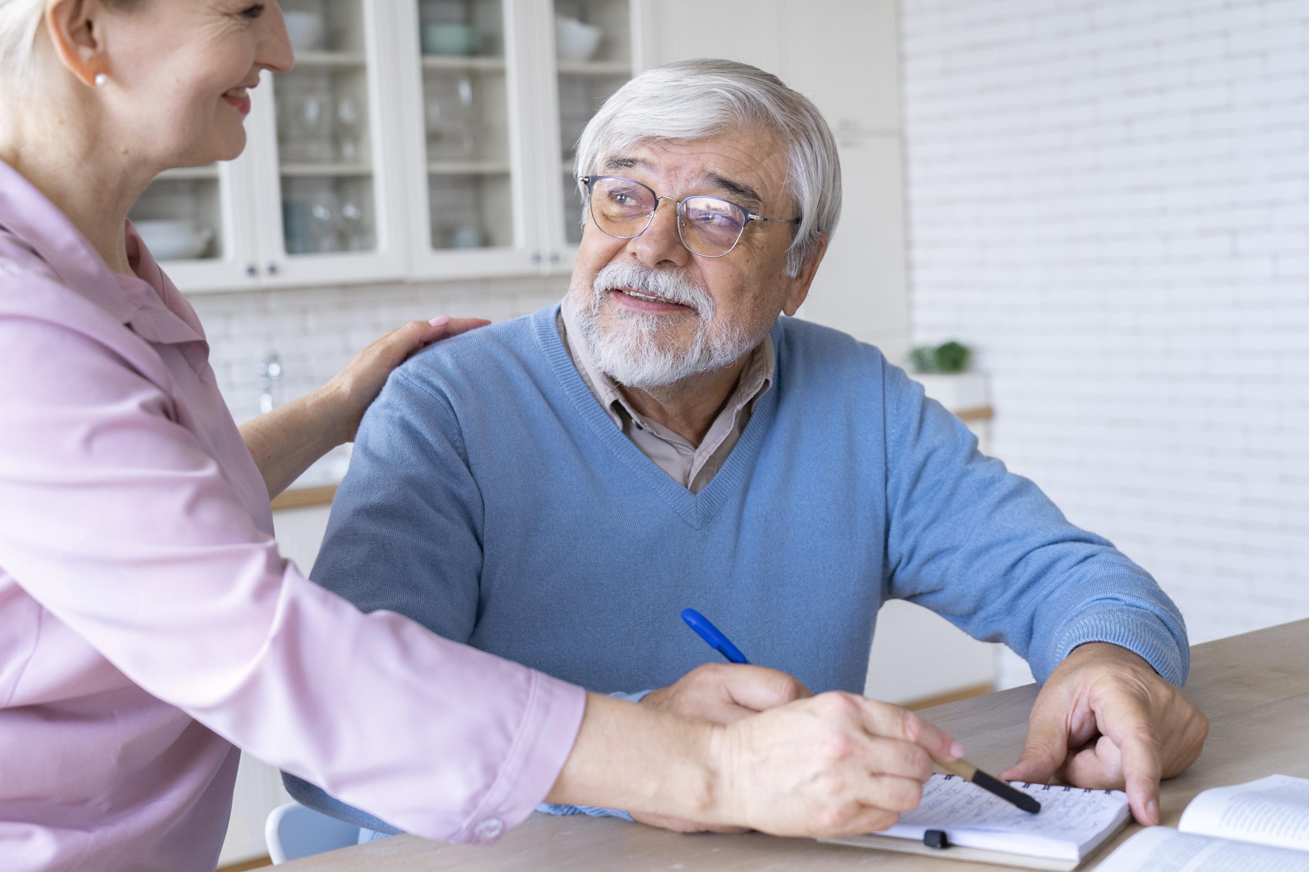 An older man with glasses writes in a notebook while a Long-Term Care Planning Attorney stands beside him, resting her hand on his shoulder in a bright kitchen.