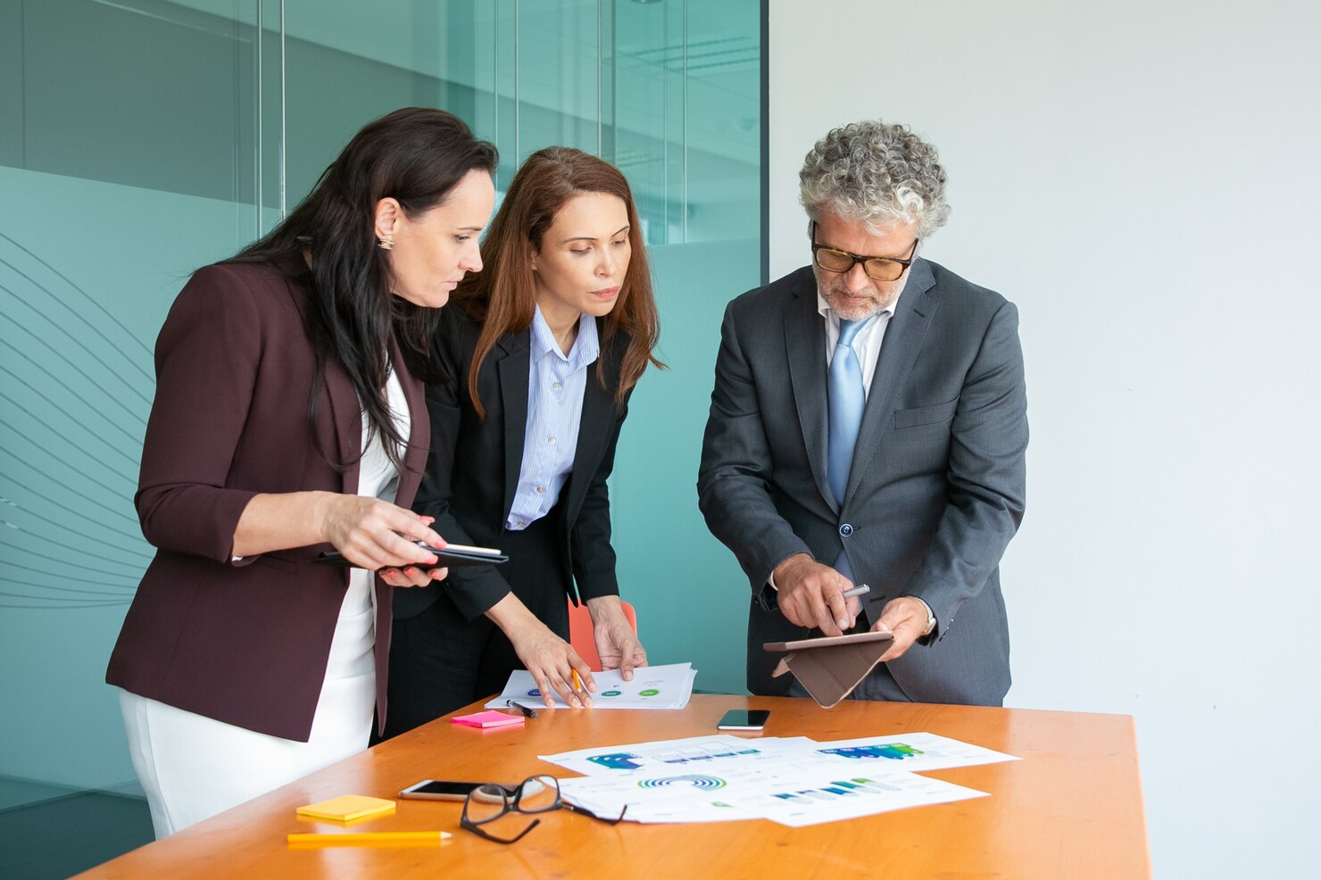 Three businesspeople in formal attire review documents and charts with a Business Formation Attorney, while one uses a tablet, in a modern office setting.