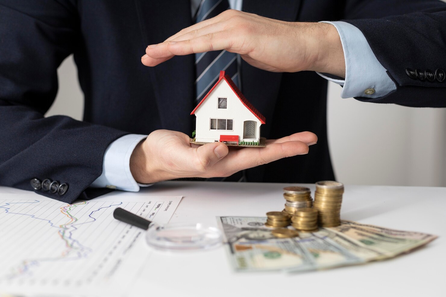 Person in a suit holds a small model house, symbolizing asset protection. Stacked coins, US dollars, financial charts, and a magnifying glass rest on the desk—like an Asset Protection Attorney at work.