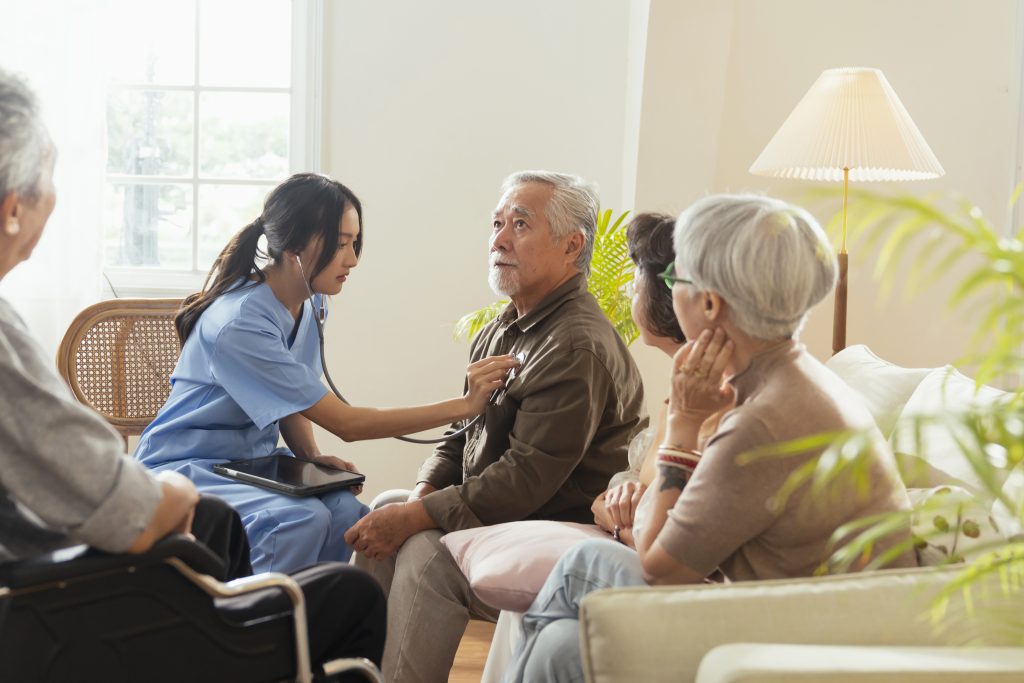 A nurse in blue scrubs checks an older man’s heart with a stethoscope while a Medicaid & Nursing Home Planning Attorney assists three other seniors nearby in a well-lit living room.