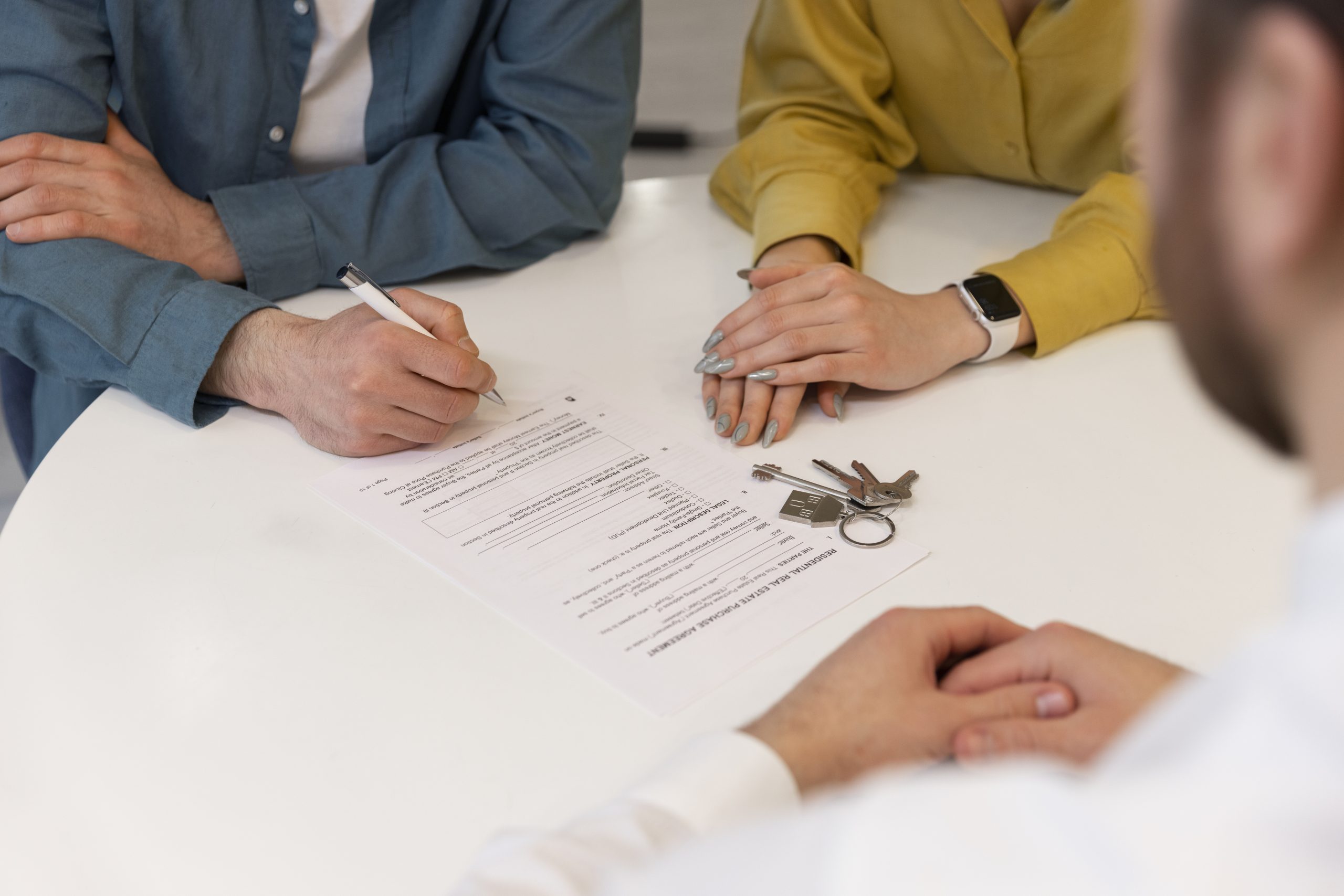 Three people sit at a table with paperwork and keys; an Advance Directive Attorney oversees as one person signs a document while another watches with hands folded.