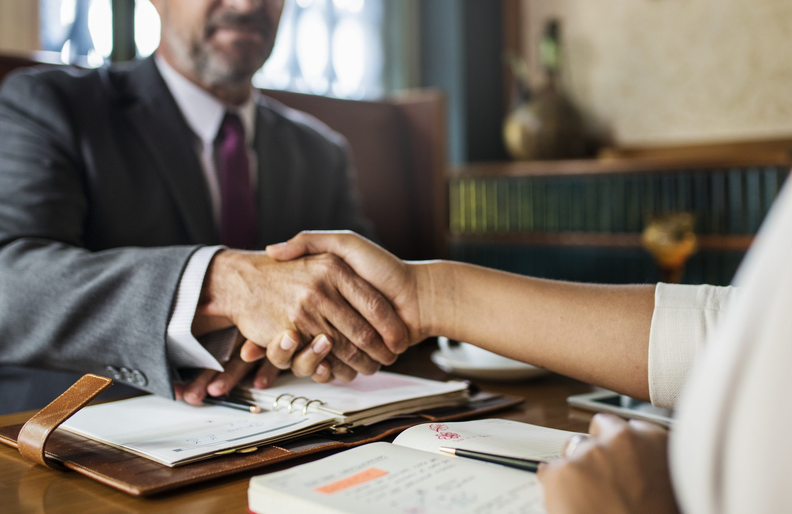 Two people are shaking hands across a desk with notebooks and documents, suggesting a business agreement or meeting with a Trust Administration Attorney.