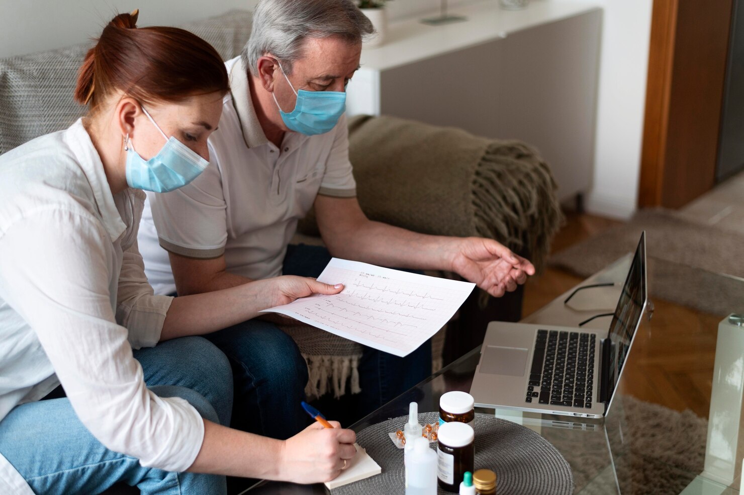 Two people wearing face masks review a medical chart and take notes while video calling about a Medicaid application, with medication bottles on the table in front of them.
