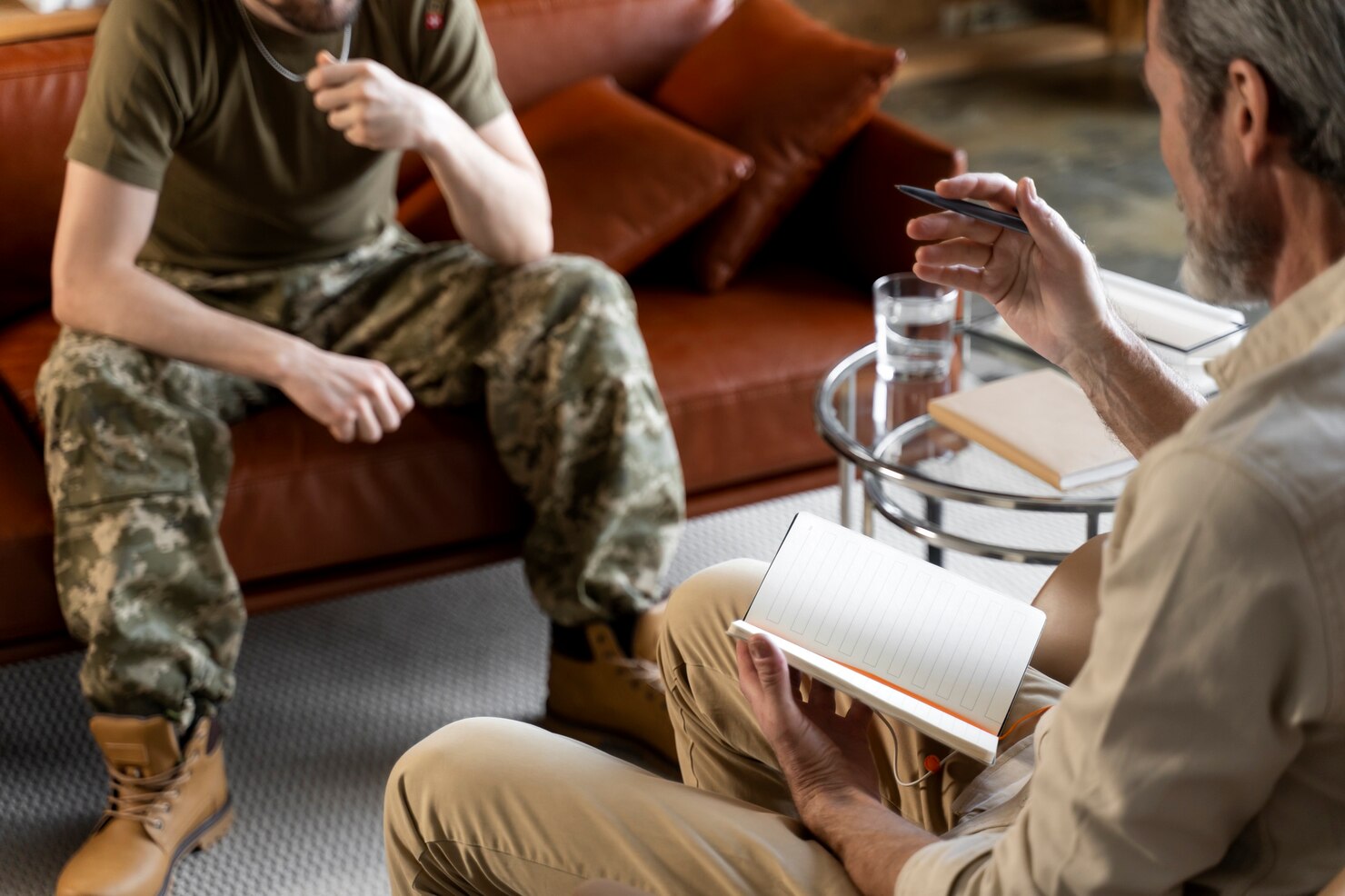 A person in military uniform sits across from another with a notebook and pen, suggesting a VA Benefits counseling session in a living room setting.