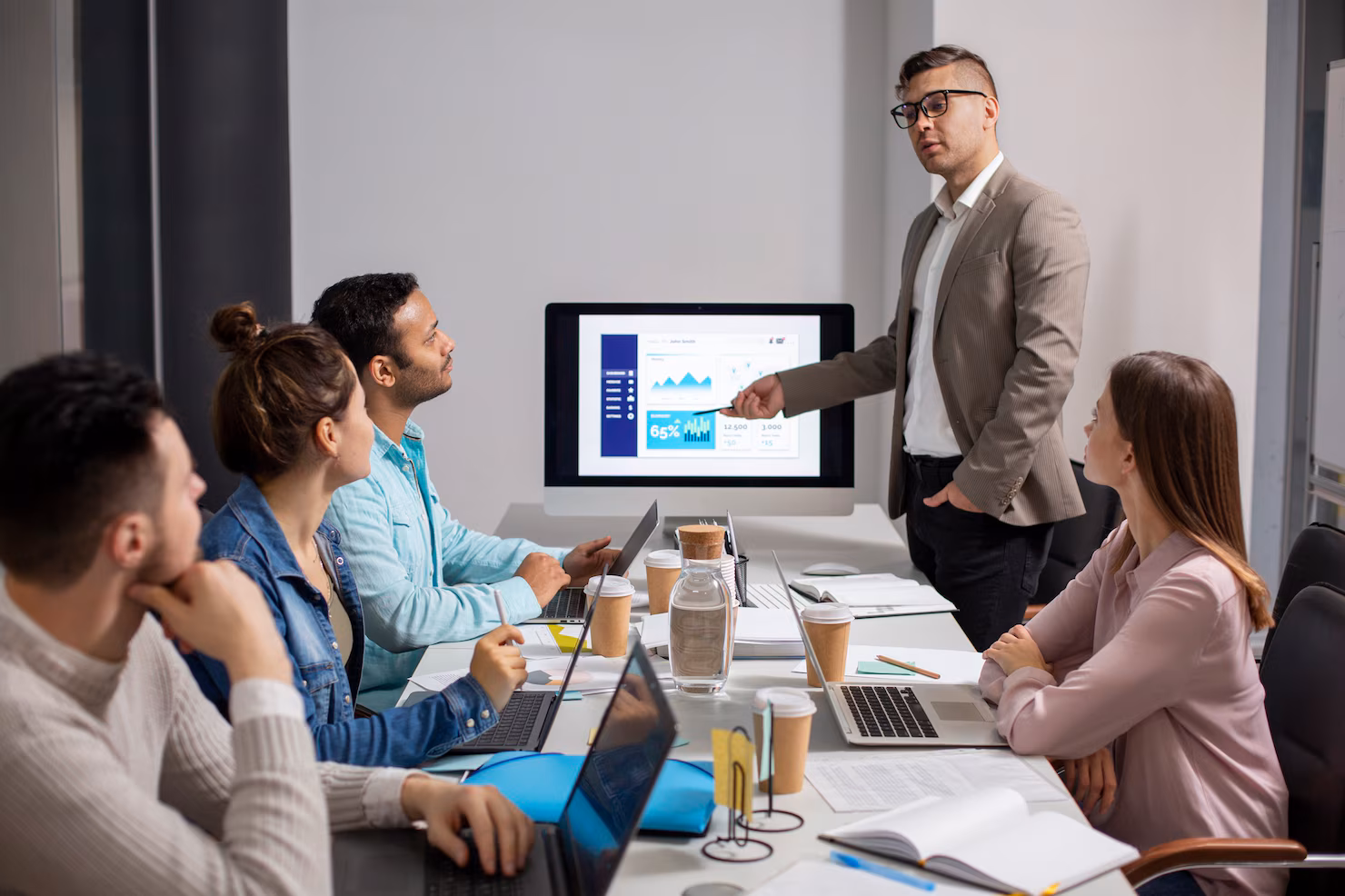A Business Formation Attorney in a blazer gives a presentation to four colleagues seated at a table with laptops, as data charts are displayed on a monitor.