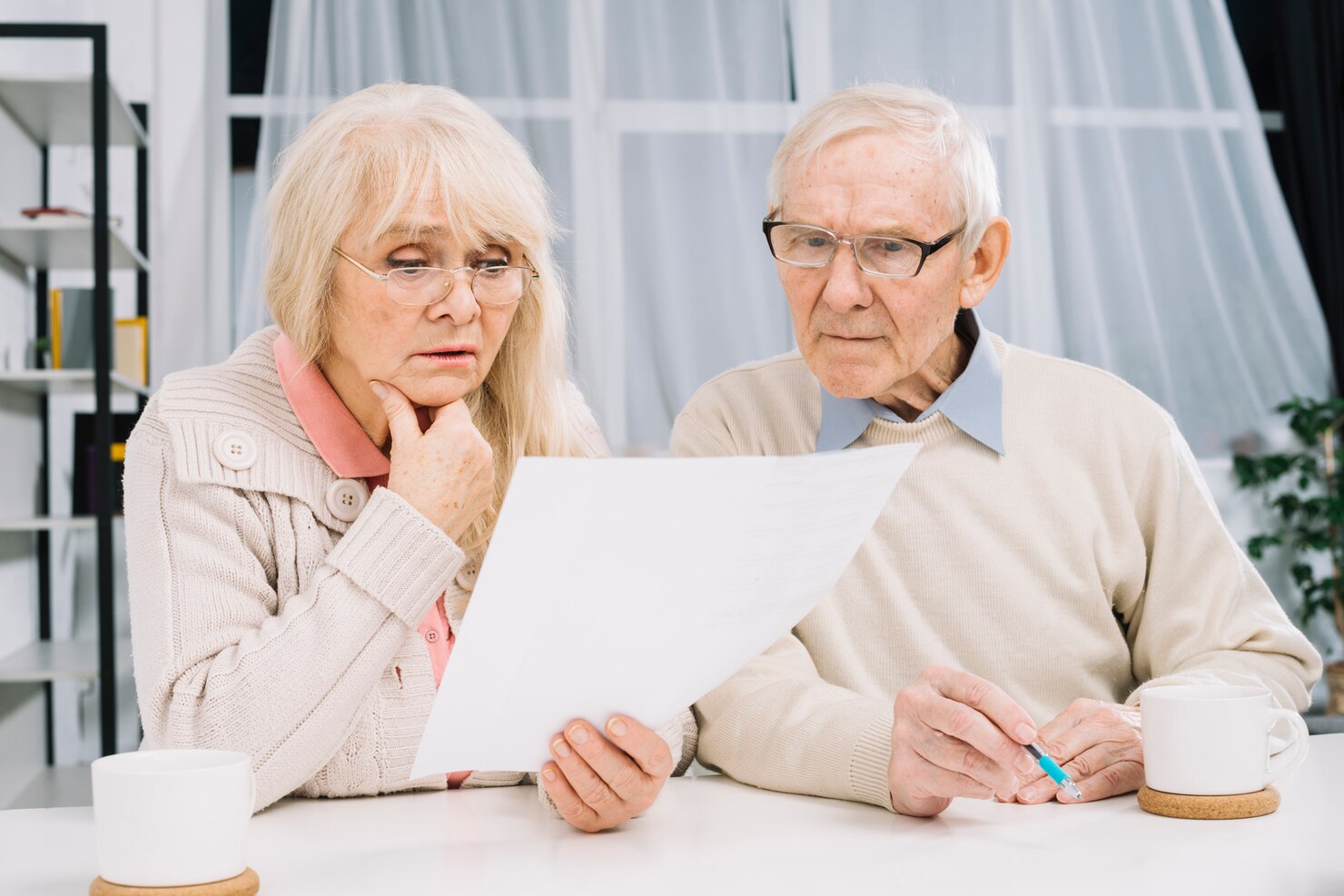 Elderly couple sitting at a table, looking intently at a sheet of paper from their Death Deed Attorney, with coffee mugs and a pen in front of them.