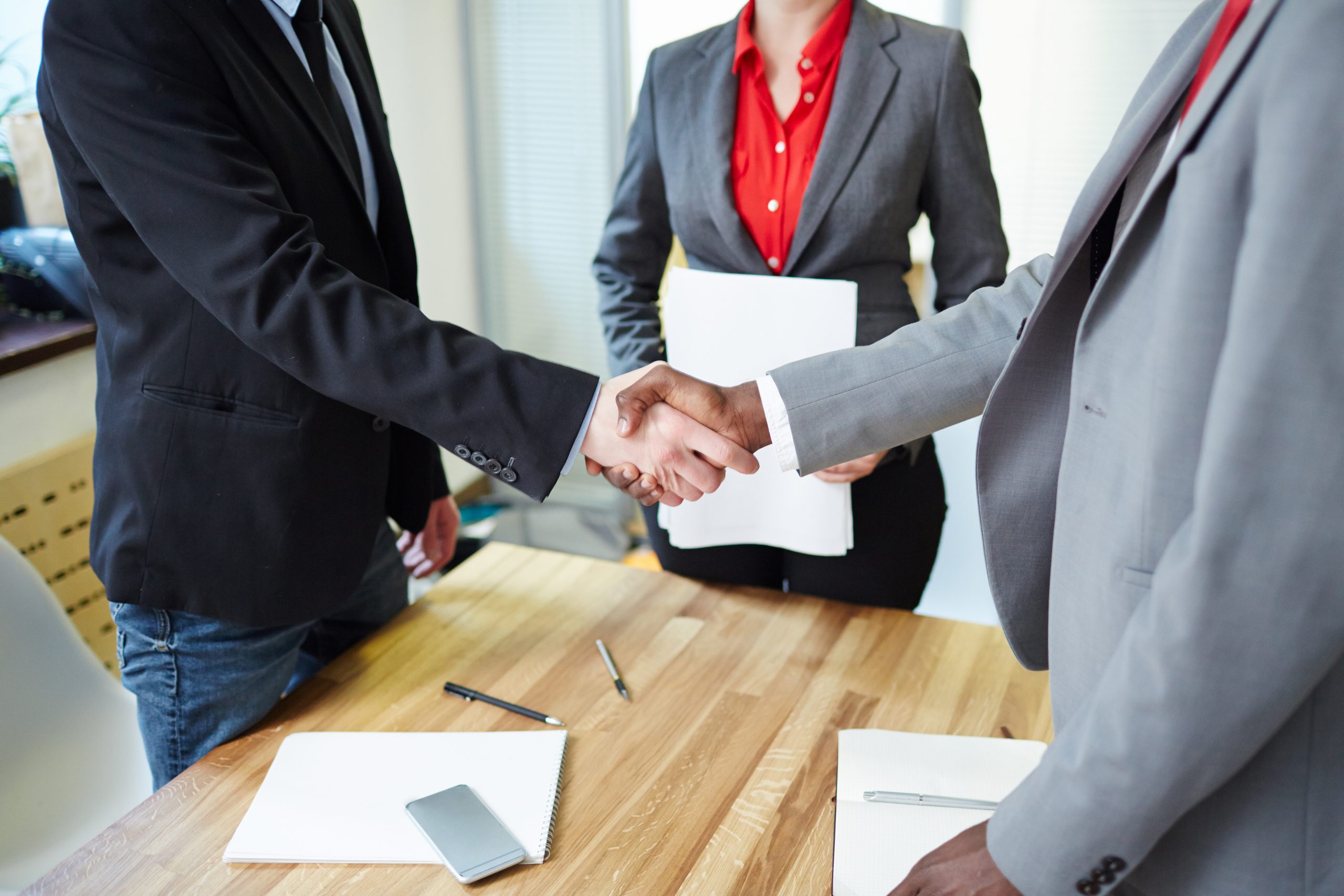 Two people in business attire shake hands across a wooden table, while a Trust Attorney stands nearby holding papers. Notebooks and pens rest on the table.