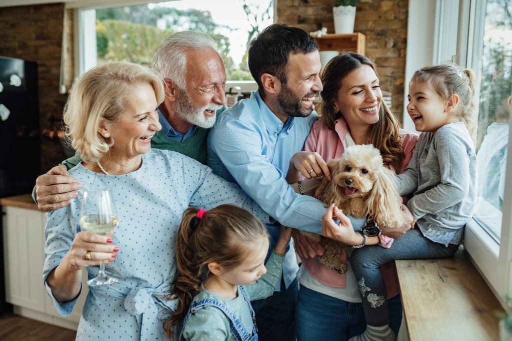 Three adults, two children, and a dog laugh together in a kitchen near a window; an Estate Planning Attorney enjoys a drink while a child pets the dog.